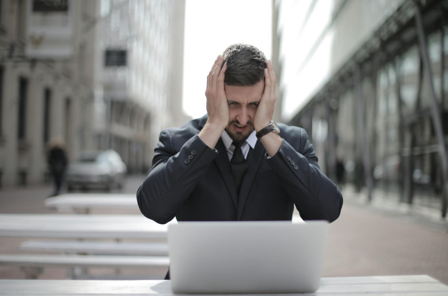 man in black suit looking at a laptop while covering his face with 2 hands